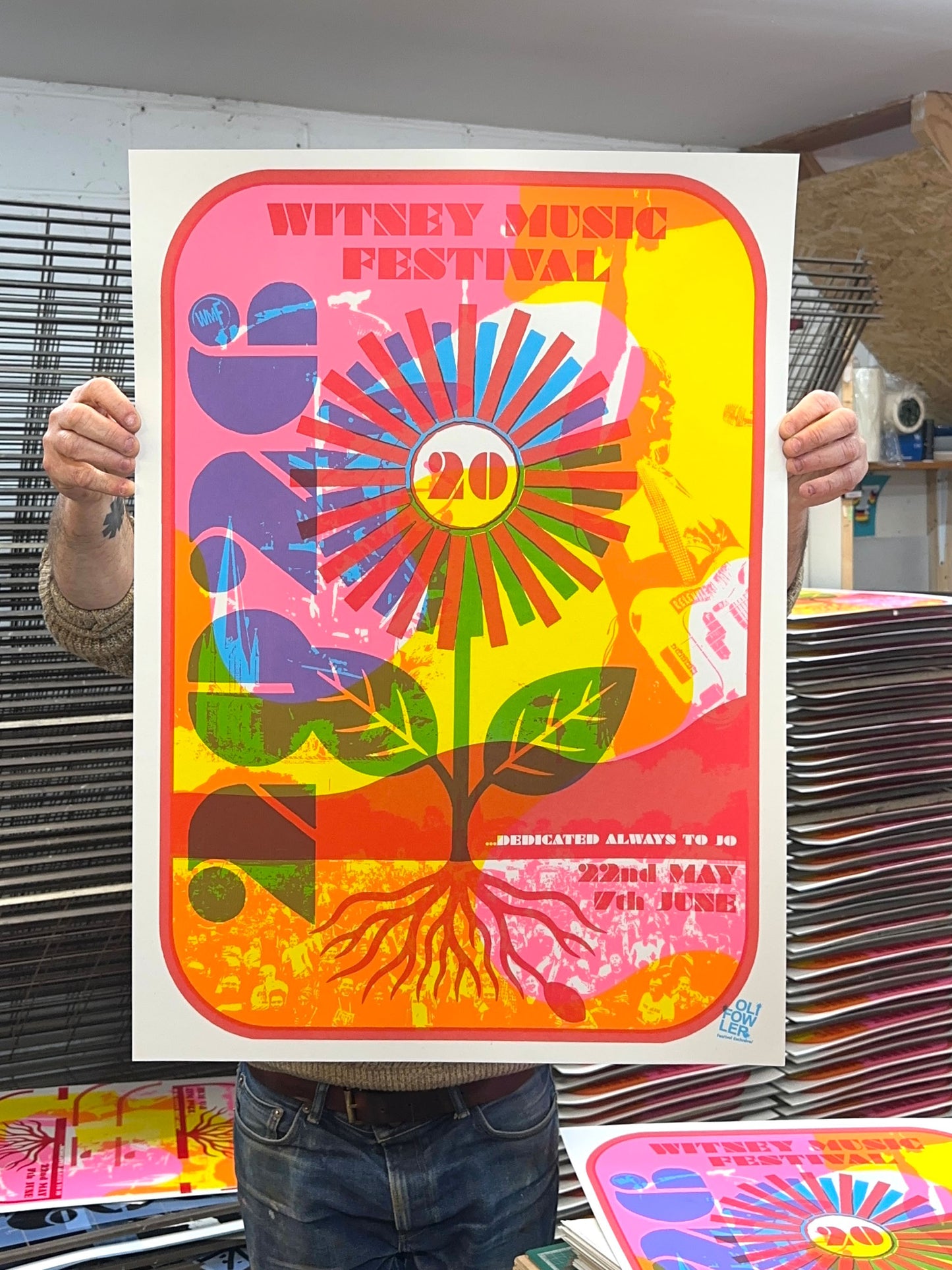 Witney Music Festival 2026 anniversary poster, hand-pulled screenprint by Oli Fowler, held up in the studio, pink and yellow flower and stem growing through a crowd scene with the number 20 at the flower's centre